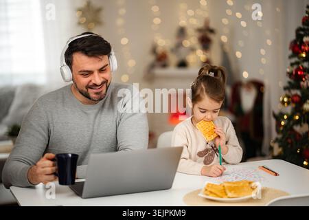 Man wearing headphones using laptop while daughter eats waffles and draws at home Stock Photo