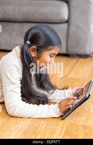 Indian child lying on hardwood floor at home using tablet stylus by gray sofa in cream-top. Girl, touchscreen, education, learning, cozy, warm, indoor Stock Photo
