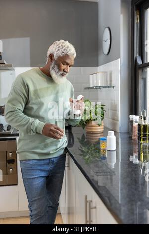 Senior African American man leaning holding white pill bottle and water glass in modern kitchen. Medication, hydration, aging, d Stock Photo