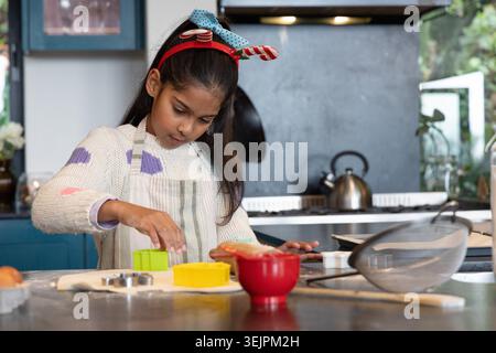 Indian girl child pressing cookie cutter into rolled dough on kitchen island wearing striped apron. Baking, pastry, home, sunlit Stock Photo