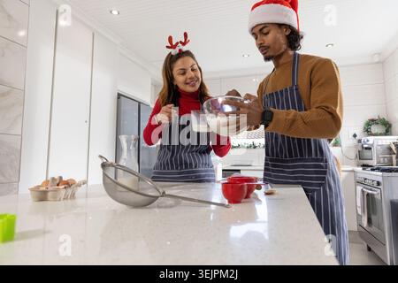 Diverse couple wearing striped aprons and festive hats mixing batter on quartz island with bowl. Kitchen, home, cooking, baking, Stock Photo