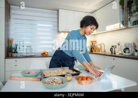 Woman cooking lasagna at home using digital tablet Stock Photo