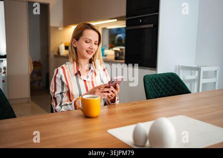 Woman drinking coffee and using smartphone in kitchen Stock Photo