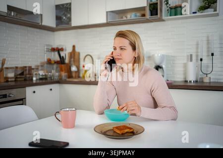 Woman talking on phone eating breakfast in kitchen Stock Photo