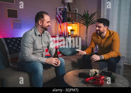 Two friends share stories and laughter over drinks in a cozy living room filled with warm ambiance and American decor Stock Photo