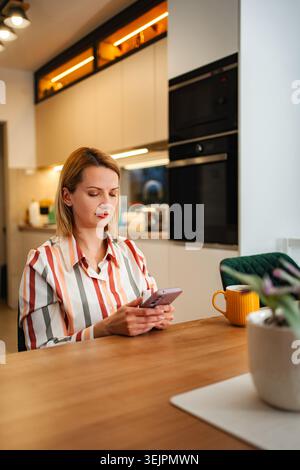 Woman using smartphone while relaxing in modern kitchen Stock Photo