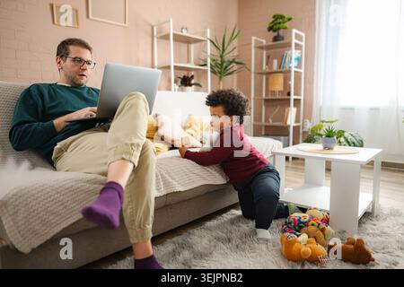 Father using laptop on sofa while his toddler son plays on the floor Stock Photo