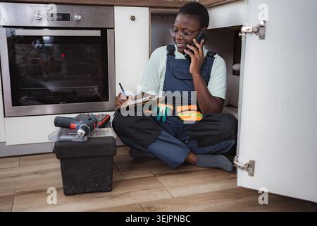 Young female plumber sitting on the floor is taking notes while speaking on the phone Stock Photo