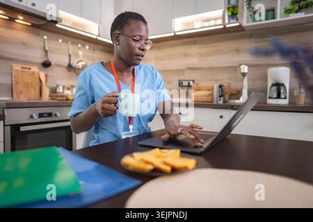 Nurse drinking coffee and working on laptop in kitchen Stock Photo