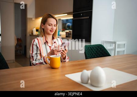 Woman using smartphone and drinking coffee in kitchen Stock Photo