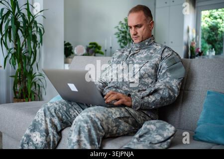Soldier in camouflage uniform using laptop on sofa at home Stock Photo