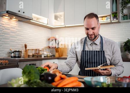 Man in apron preparing fresh vegetables following recipe book Stock Photo