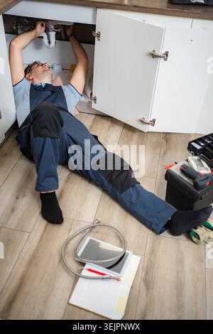 Plumber lying on floor repairing kitchen sink drainage system Stock Photo