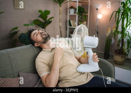 Man relaxing on sofa holding electric fan trying to refresh during summer heat wave at home Stock Photo