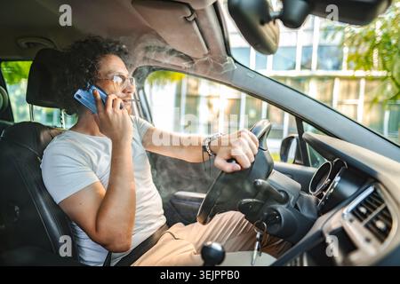 Man with curly hair driving a car, smoking a cigarette and using phone Stock Photo