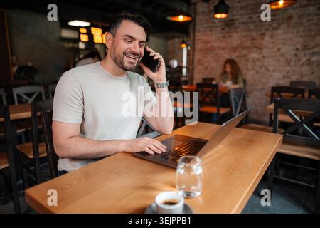 Happy businessman working remotely from a cafe, using laptop and talking on the phone Stock Photo