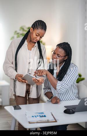 Two black business women collaborating on a digital tablet in a modern office environment Stock Photo