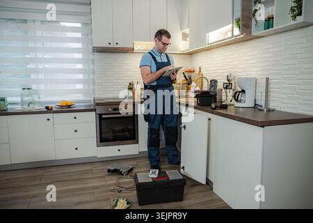 Plumber in overalls using tablet while repairing kitchen sink Stock Photo