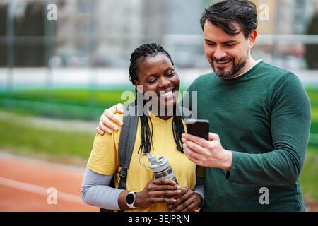 Diverse couple smiling using smartphone on running track Stock Photo