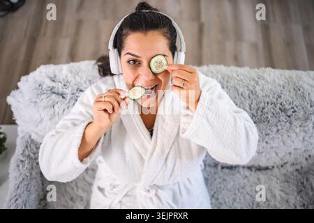 Mid adult woman wearing bathrobe is listening to music and applying cucumber slices on her eyes and mouth Stock Photo