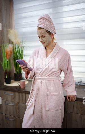 Young woman wearing bathrobe and face mask checking her phone in the kitchen Stock Photo