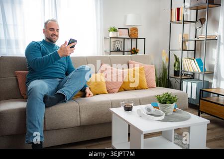 Smiling man enjoying smartphone while relaxing on sofa at home Stock Photo