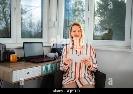 Woman smiling using digital tablet next to home office desk Stock Photo