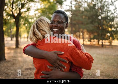 Two happy multi-ethnic friends hugging each other in a park Stock Photo