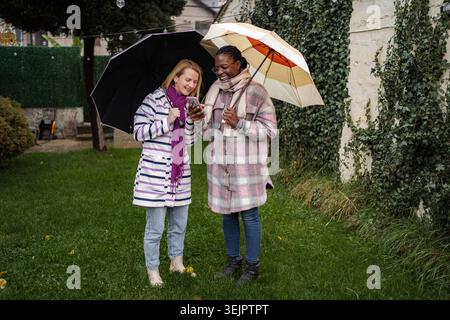 Two diverse women standing in a yard using a smartphone under umbrellas in the rain Stock Photo