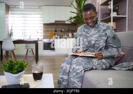 Smiling african american female soldier is sitting on the sofa at home, enjoying a relaxing moment reading a book Stock Photo