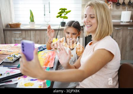 Mother and daughter engaged in a cheerful video call while painting together at home in a cozy kitchen setting Stock Photo