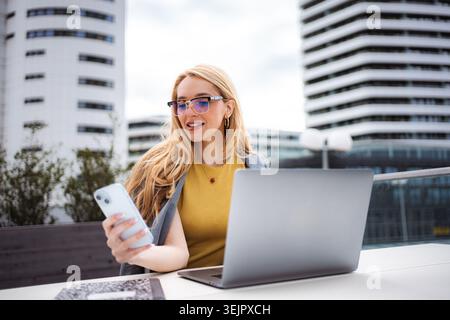 Businesswoman checking smartphone while working on laptop outdoors Stock Photo
