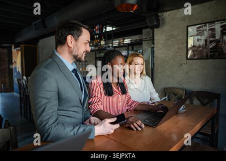 Three diverse colleagues discussing business strategy and using a laptop in a cafe Stock Photo