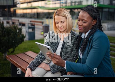Two businesswomen collaborating on a digital tablet in a park setting Stock Photo