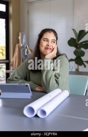 Indian woman sitting at work table, resting cheek on hand, reviewing tablet and rolled blueprints. Studio, architecture, plans, Stock Photo