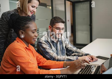 Three individuals are seated around a table, focused on a laptop screen in front of them. They appear engaged in a discussion or collaborative work, w Stock Photo
