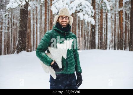 Cheerful bearded male wears warm anorak and hat, walks in winter forest, holds artificial fir tree, has festive mood, going to c Stock Photo