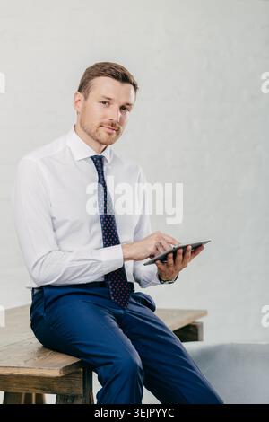 Serious male entrepreneur watches video on digital tablet, messages with business partners, dressed elegantly, sits on wooden table, poses against whi Stock Photo
