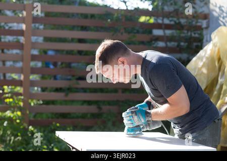 The man carpenter is engaged in steel work near the house in the summer. a man using wood polishing machine grinding sanding woo Stock Photo