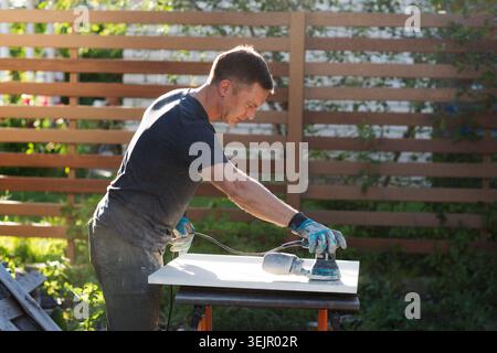 The man carpenter is engaged in steel work near the house in the summer. a man using wood polishing machine grinding sanding woo Stock Photo