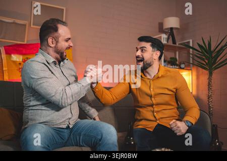 Two friends celebrate in a cozy living room while enjoying a friendly arm wrestling match during a festive gathering Stock Photo