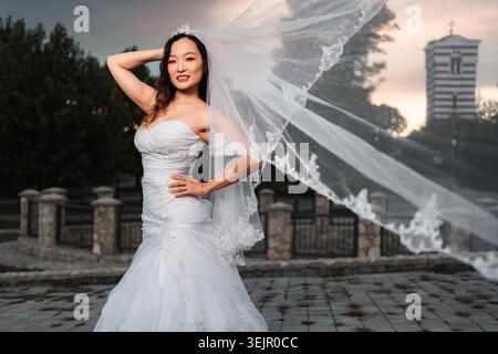 Smiling bride posing outdoors, her white veil blowing in the wind Stock Photo