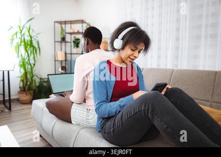 Two sisters are sitting back to back on the sofa, one is working on a laptop and the other is listening to music and using her smartphone photo Stock Photo