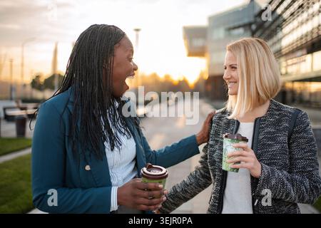 Diverse women colleagues laughing drinking coffee outdoors Stock Photo