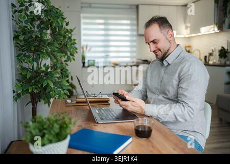 Smiling man working from home using smartphone and laptop Stock Photo