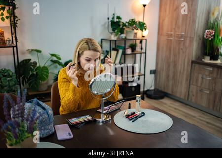 Woman applying makeup in a cozy indoor setting with plants and warm lighting during afternoon hours Stock Photo