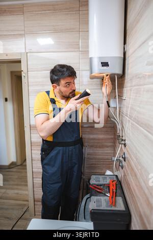 Plumber inspecting electric water heater using flashlight in bathroom Stock Photo