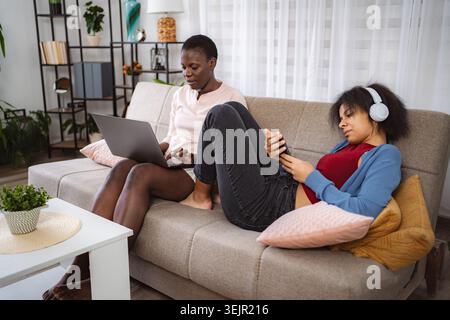 Two Young Women Relaxing on Sofa, Using Laptop and Smartphone Stock Photo