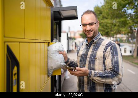 Man using a package delivery locker on a sunny day in an urban setting while checking his phone Stock Photo