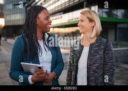 Diverse businesswomen smiling and talking while walking in city Stock Photo